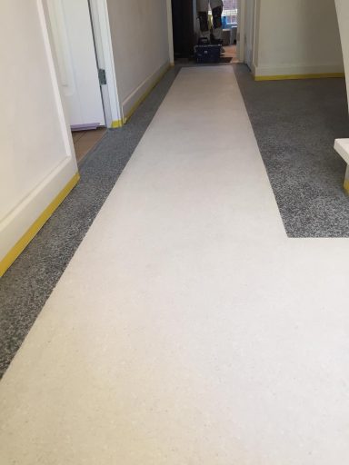 Lange gang met een lichte vloerbedekking en gele plinten aan de zijkanten.